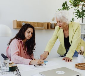 A senior woman in a yellow blazer leans over a desk to guide a younger colleague who is reviewing documents in a bright, modern office.