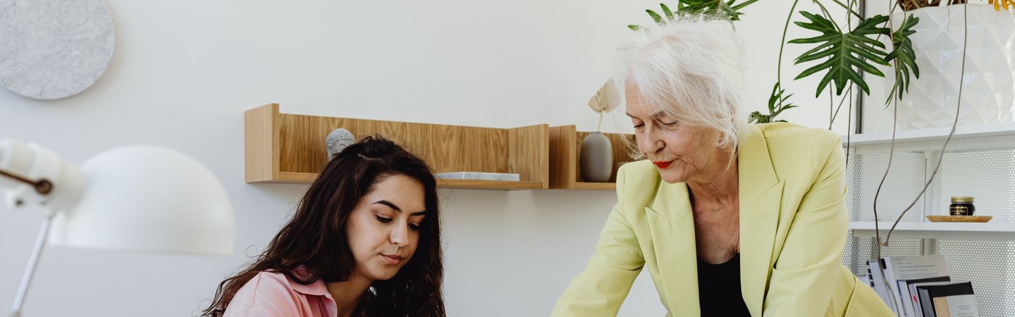 A senior woman in a yellow blazer leans over a desk to guide a younger colleague who is reviewing documents in a bright, modern office.