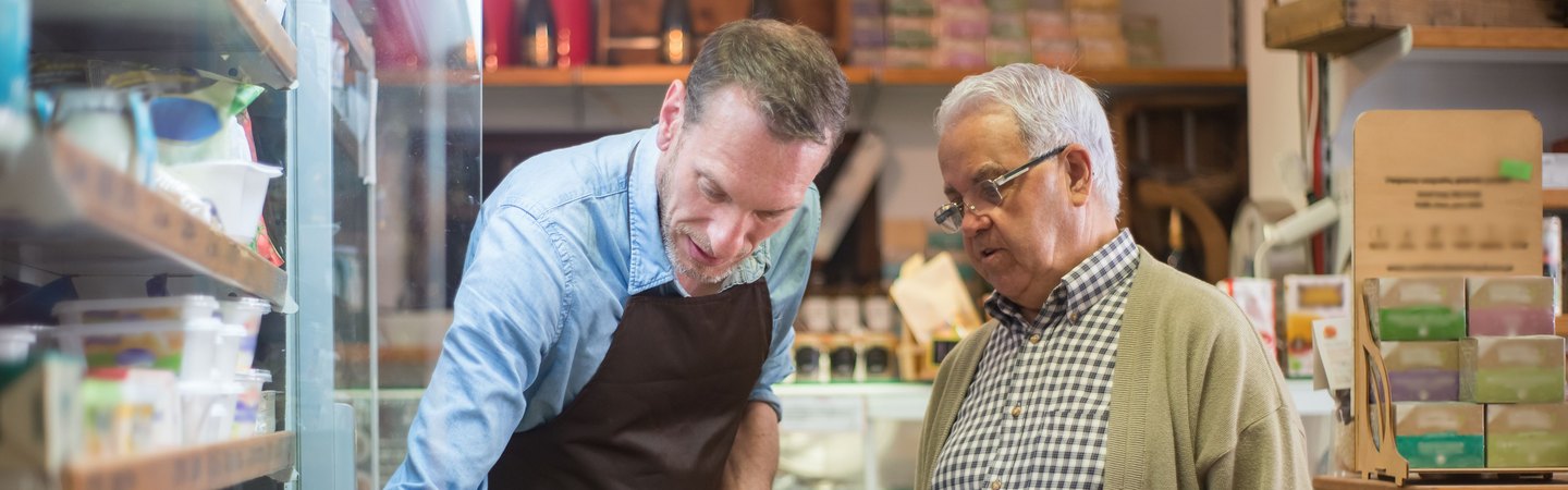 A shop worker in an apron helping an older customer at a grocery counter