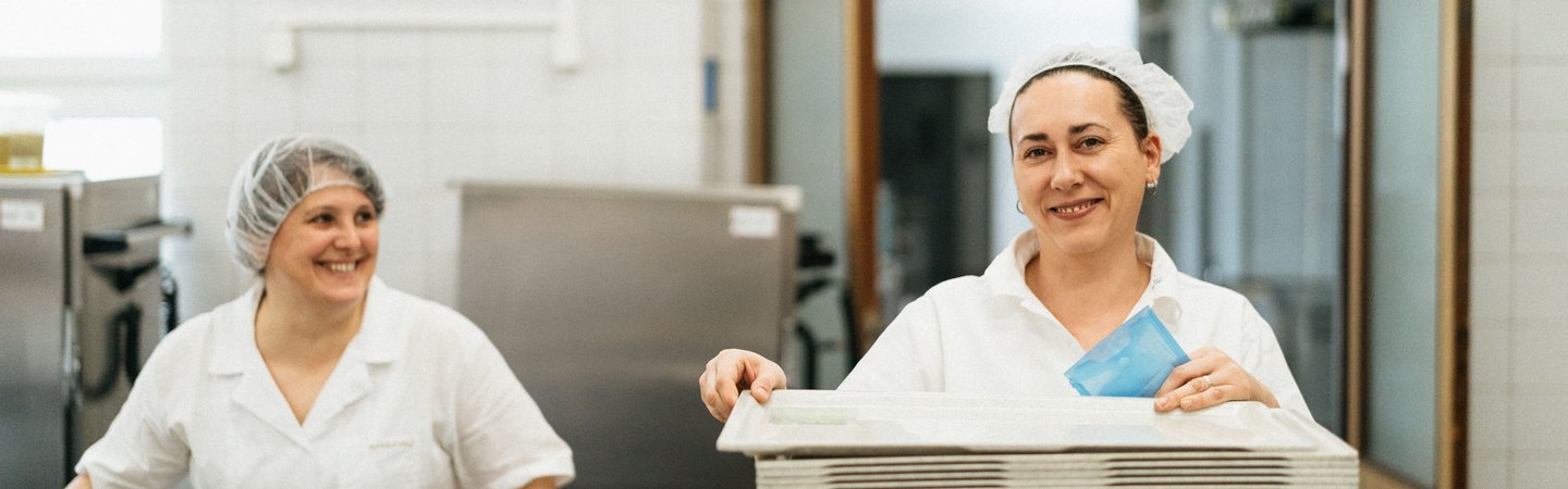 Two smiling women in white uniforms and hair nets work together in a bright commercial kitchen, stacking trays at a prep station.