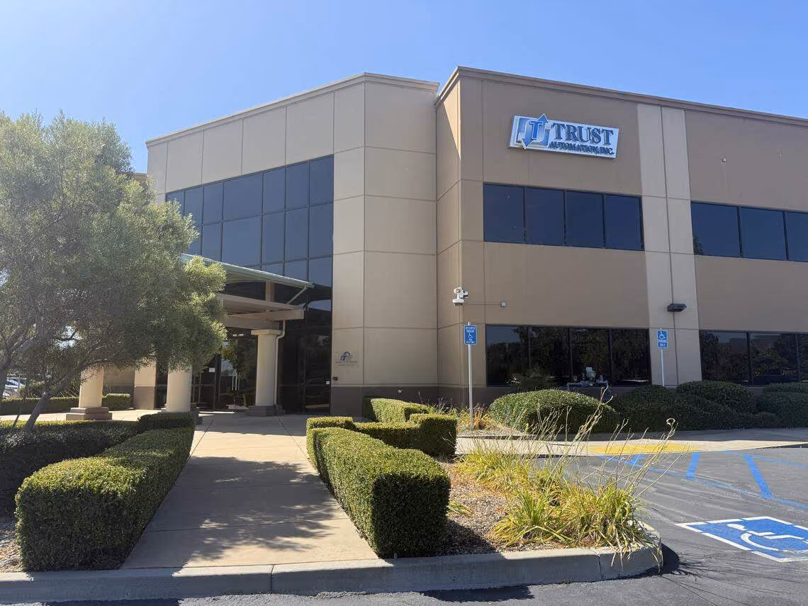 Modern two-story office building with beige exterior and dark-tinted windows labeled Trust Automation Inc.
