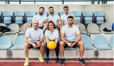 Sechs Personen in weißem T-Shirt sitzen auf blauen Sitzen in einem Stadion, eine Frau in der Mitte hält einen gelben Ball.