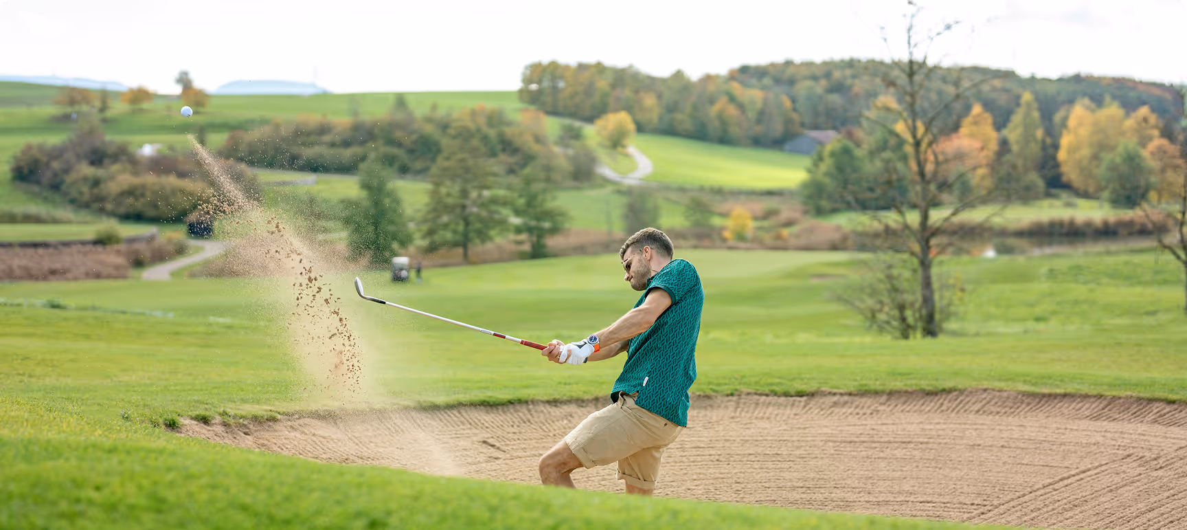 Mann schlägt einen Golfball aus einem Sandbunker auf einem grünen Golfplatz mit Bäumen und Hügeln im Hintergrund.