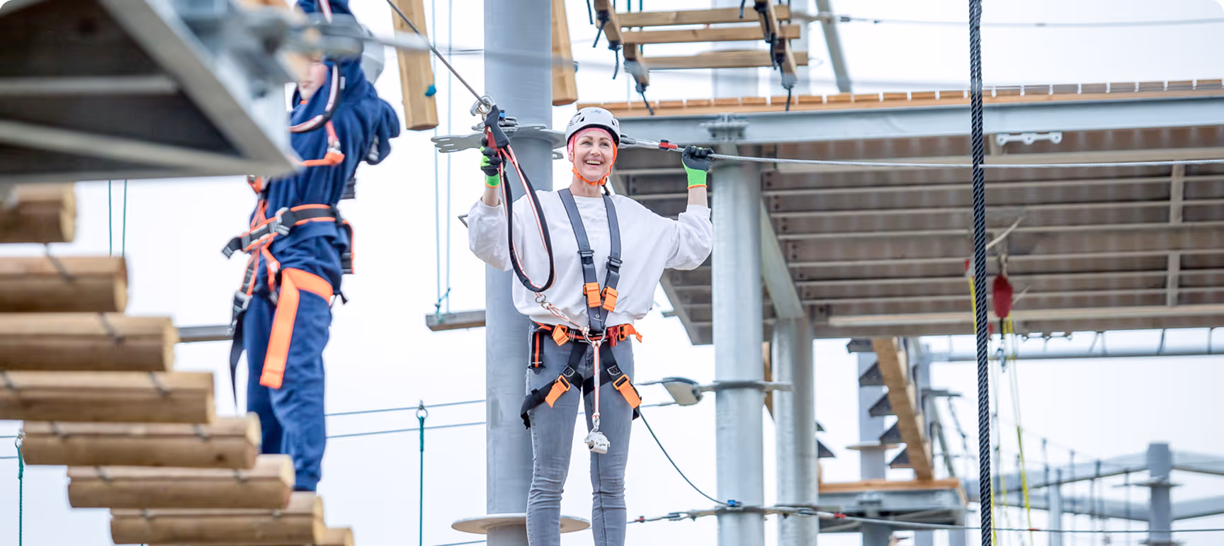 Frau mit Helm und Kletterausrüstung beim Hochseilgarten, lächelnd an Kabelsicherung haltend.