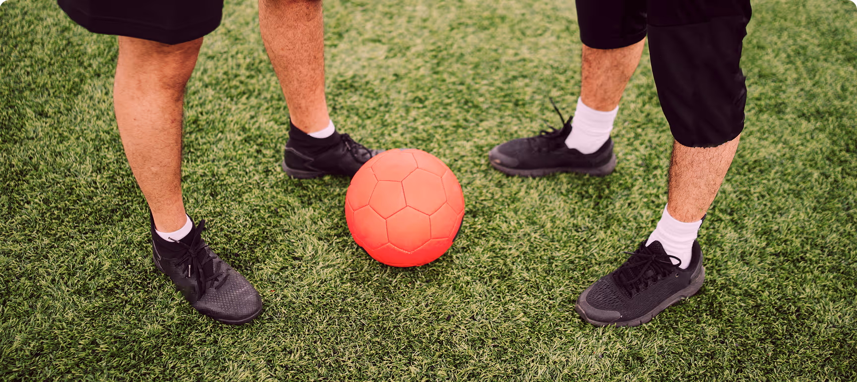 Beine von zwei Personen mit schwarzen Sportschuhen und weißen Socken stehen auf grünem Kunstrasen um einen roten Fußball.