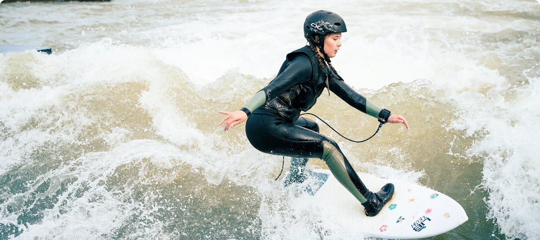 Frau surft auf einem weißen Surfbrett mit Blumenmuster auf einer Welle, trägt schwarzen Neoprenanzug und Helm.