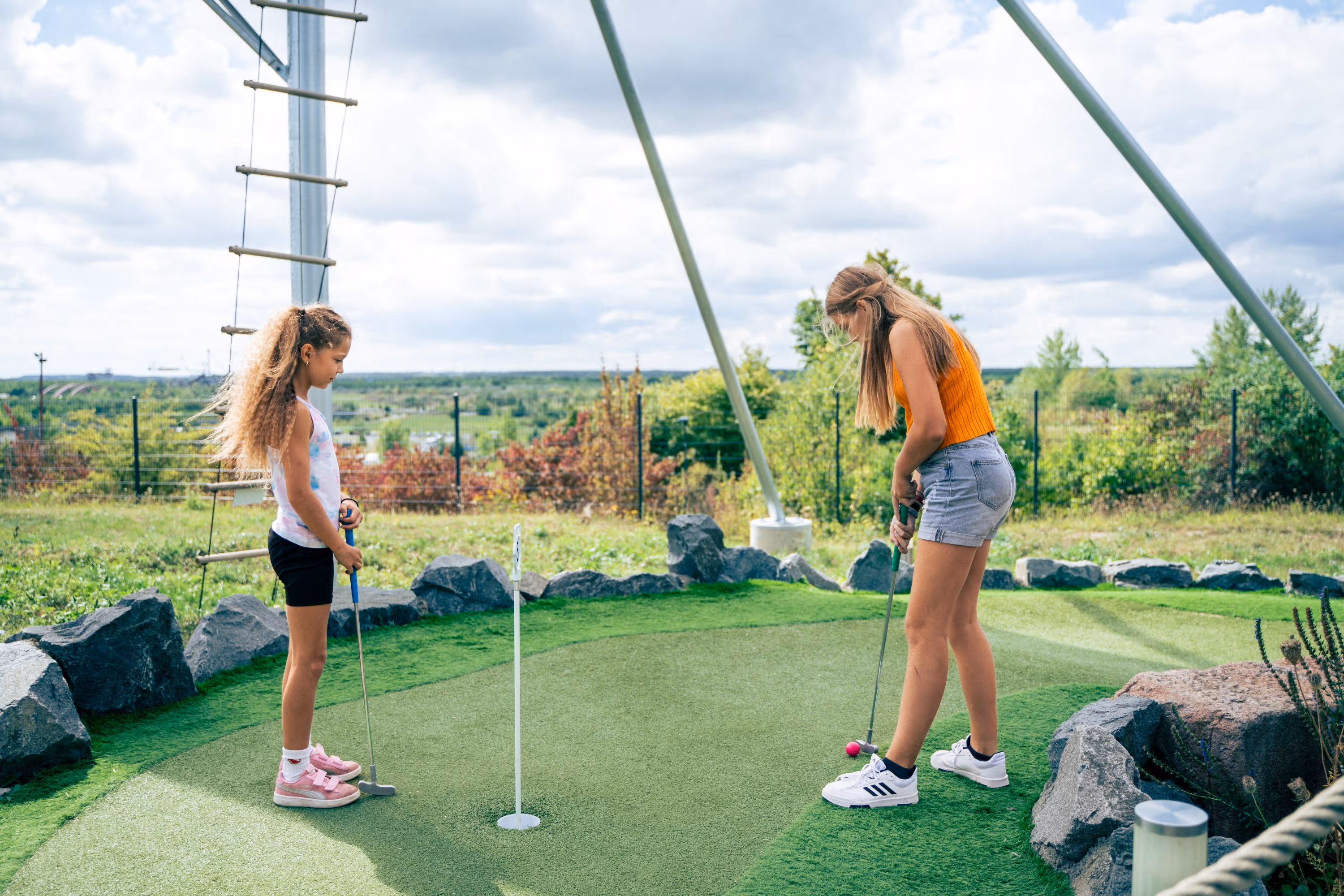 Zwei Mädchen spielen Minigolf auf einem Außengelände mit künstlichem Gras und Felsen.