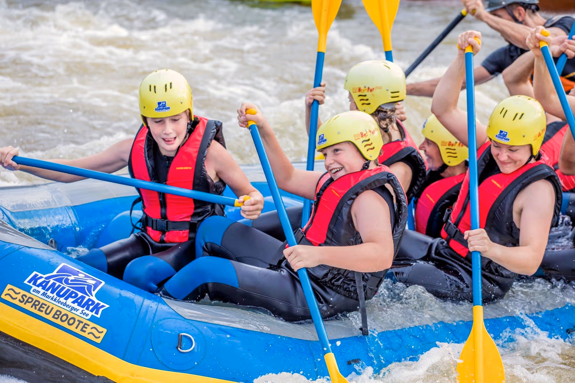 Gruppe von Kindern mit gelben Helmen und roten Schwimmwesten, die in einem blauen Schlauchboot auf einem Wildwasserfluss paddeln.