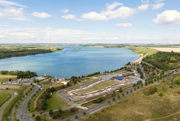 Luftaufnahme eines großen Sees mit umliegenden Grünflächen, Straßen und einem Baugebiet.