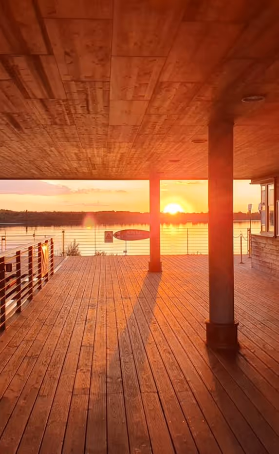 Holzterrasse mit Überdachung und Säulen, Sonnenuntergang über einem ruhigen See im Hintergrund.