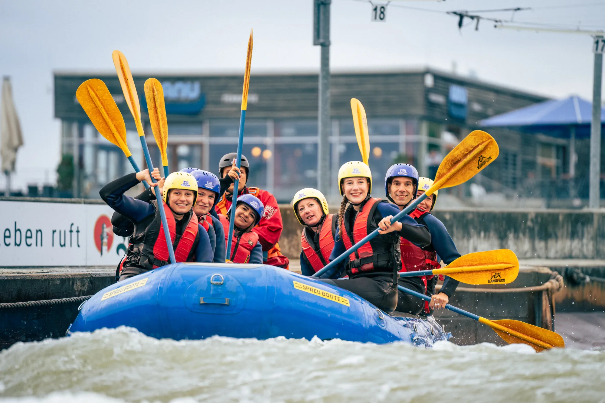 Gruppe von sieben Personen mit Helmen und roten Schwimmwesten beim Rafting im blauen Schlauchboot mit gelben Paddeln.