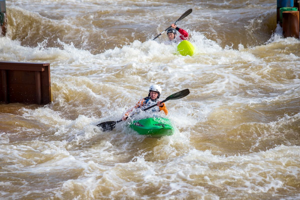 Zwei Personen in gelben Kajaks paddeln in einem Wildwasserkanal, eine lächelt in die Kamera.
