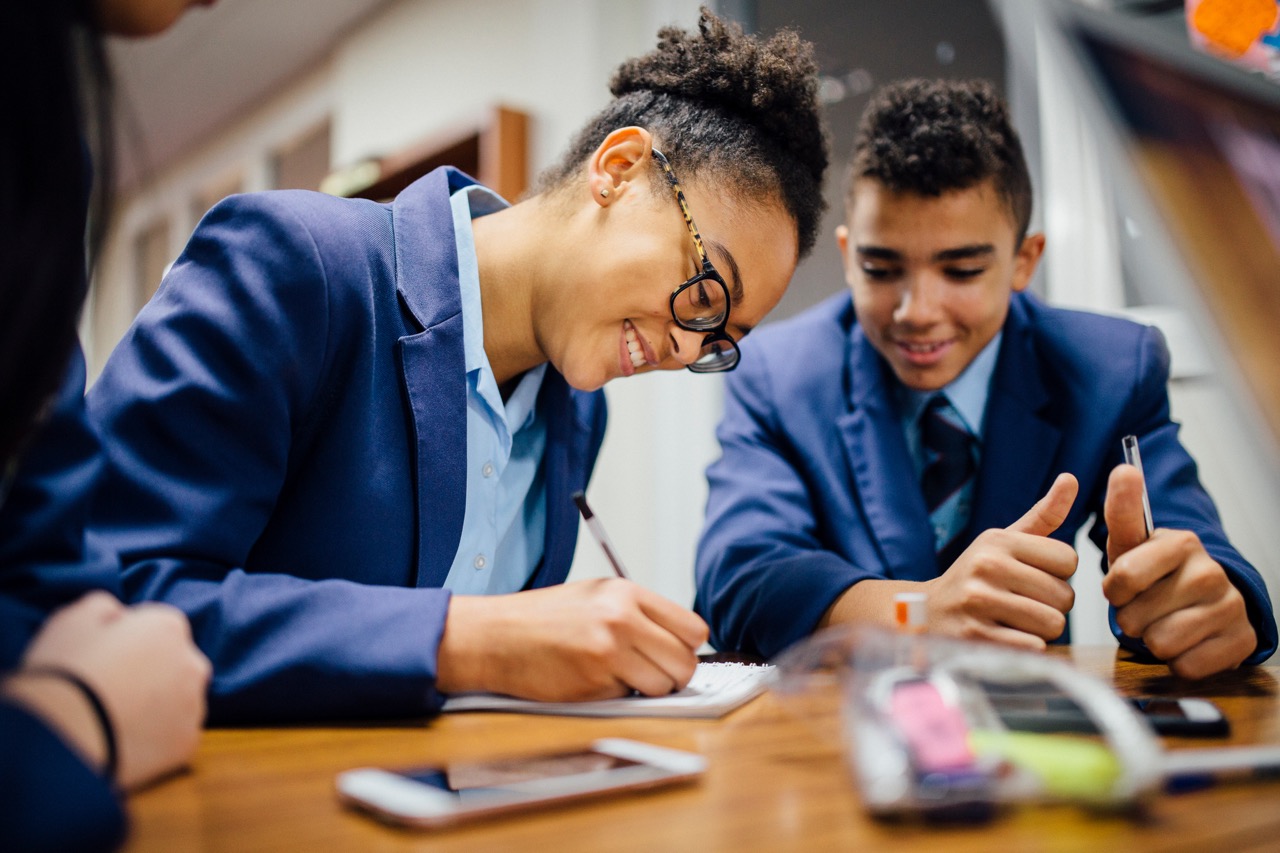 Two students in blue uniforms working together at a desk, one writing and the other giving a thumbs-up.
