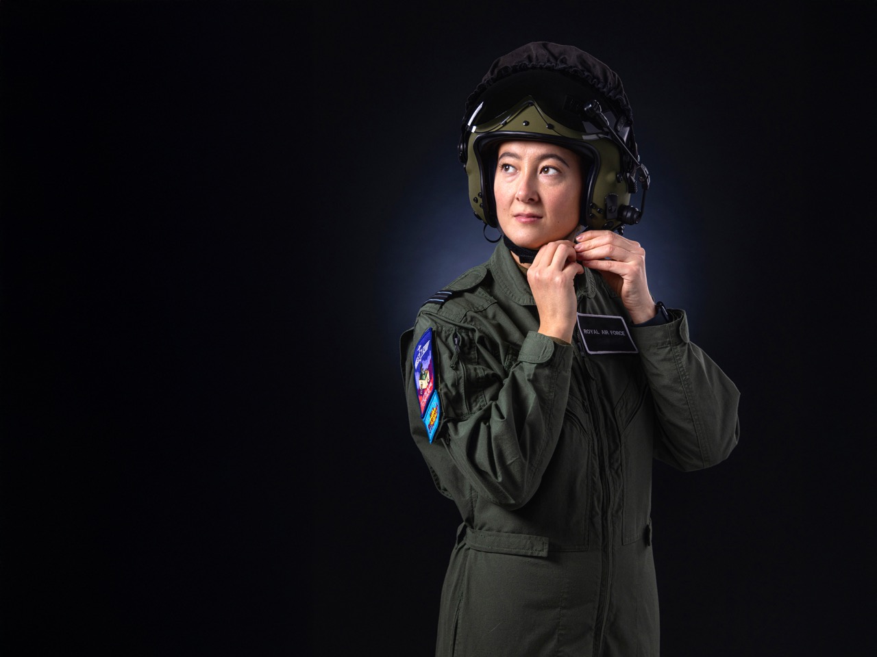 Woman in a Royal Air Force flight suit adjusting her helmet against a dark background.