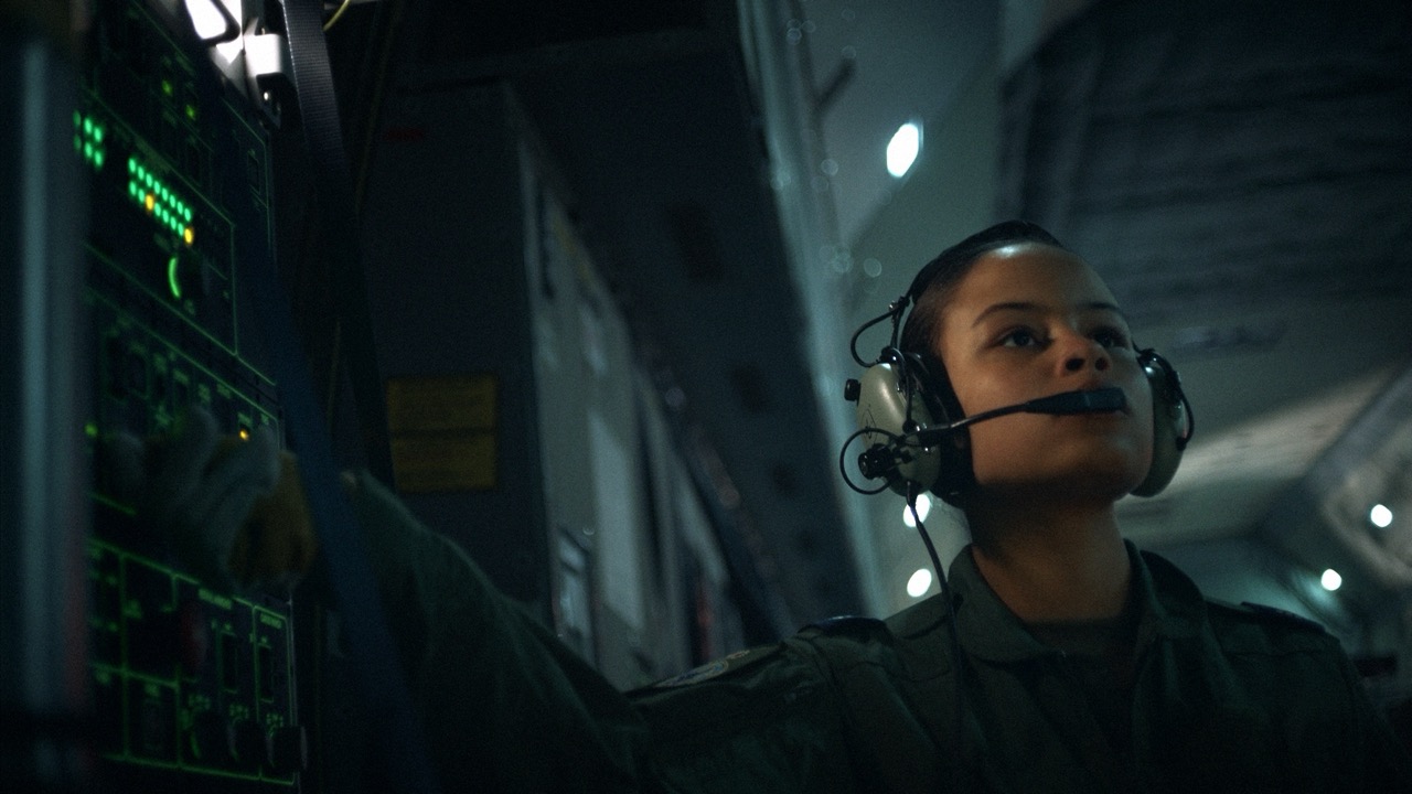 Military personnel wearing a headset and uniform operating control panel in a dimly lit aircraft interior.