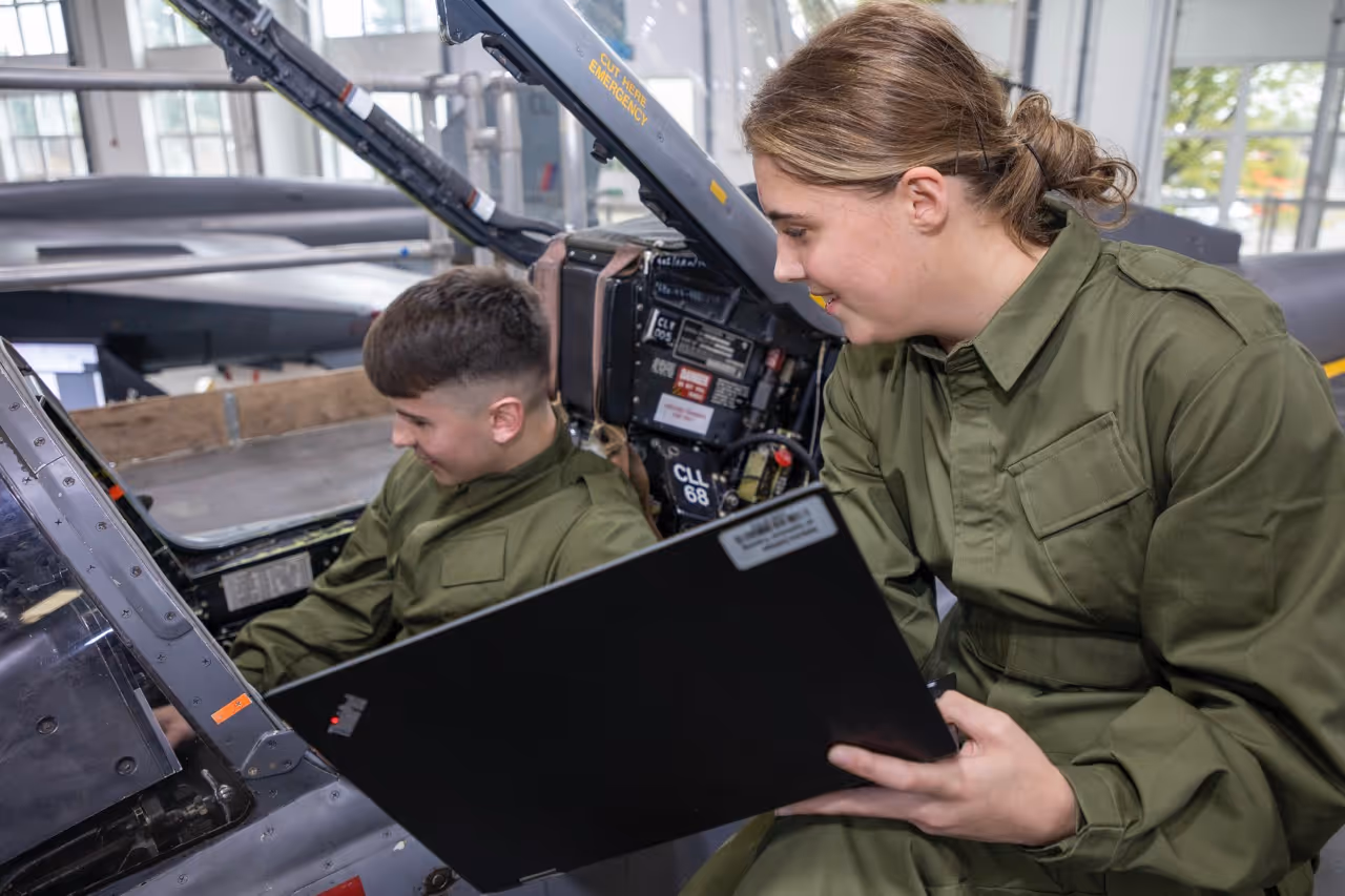 Two people in military green uniforms examining cockpit controls, one holding a laptop.