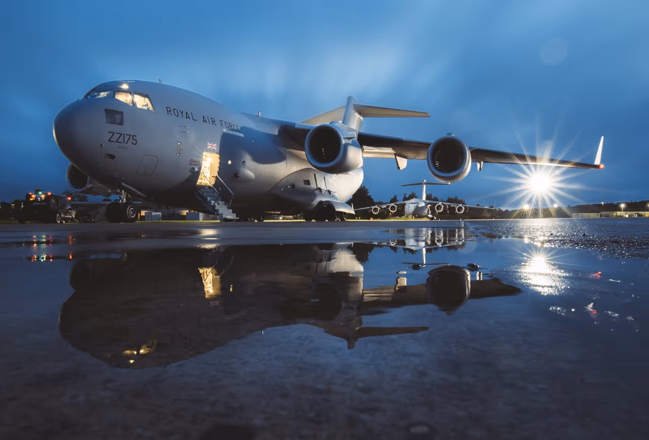 Royal Air Force military cargo planes parked on wet tarmac at dusk with reflections and bright runway lights.