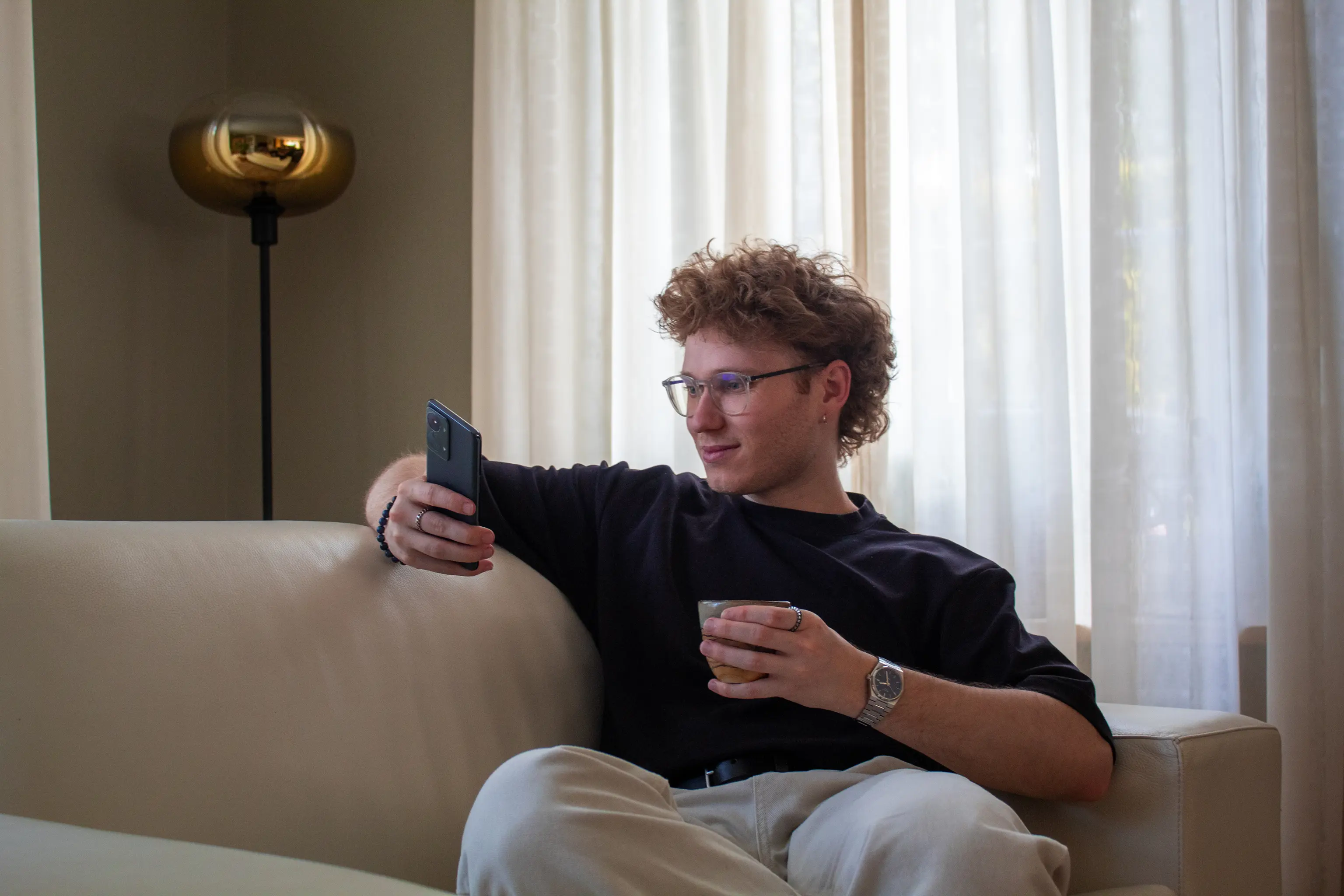 A young man with glasses sitting on a couch, looking at his phone and holding a cup.