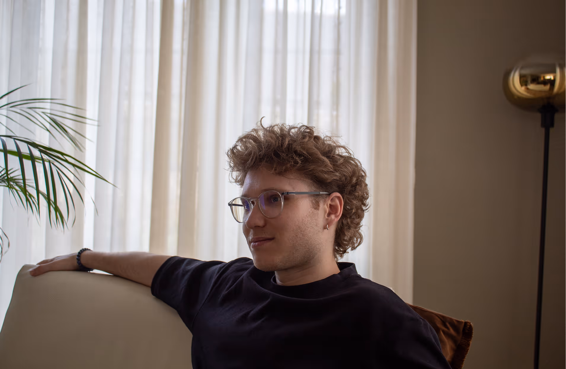 Young man with curly hair and glasses sitting on a couch by sheer curtains indoors.