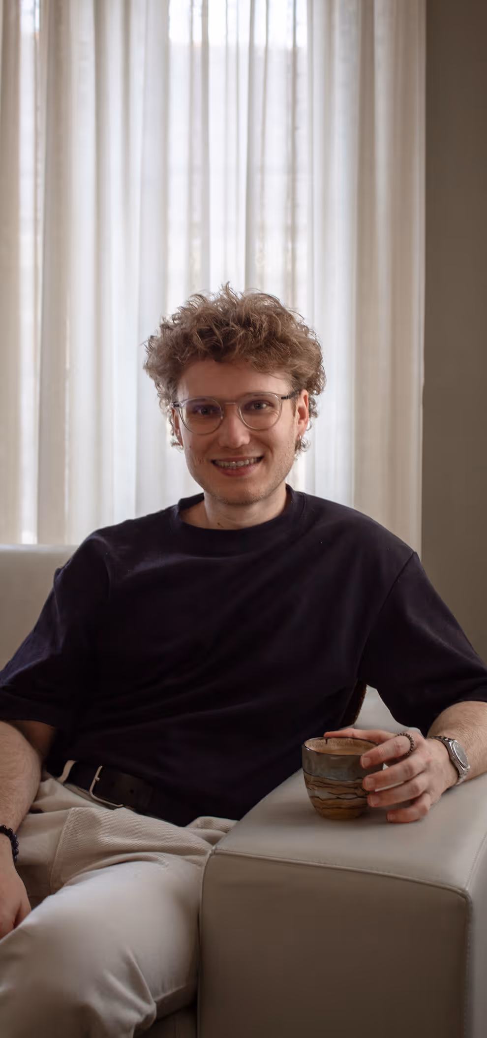 Young man with curly hair and glasses sitting on a couch holding a cup, smiling at the camera.