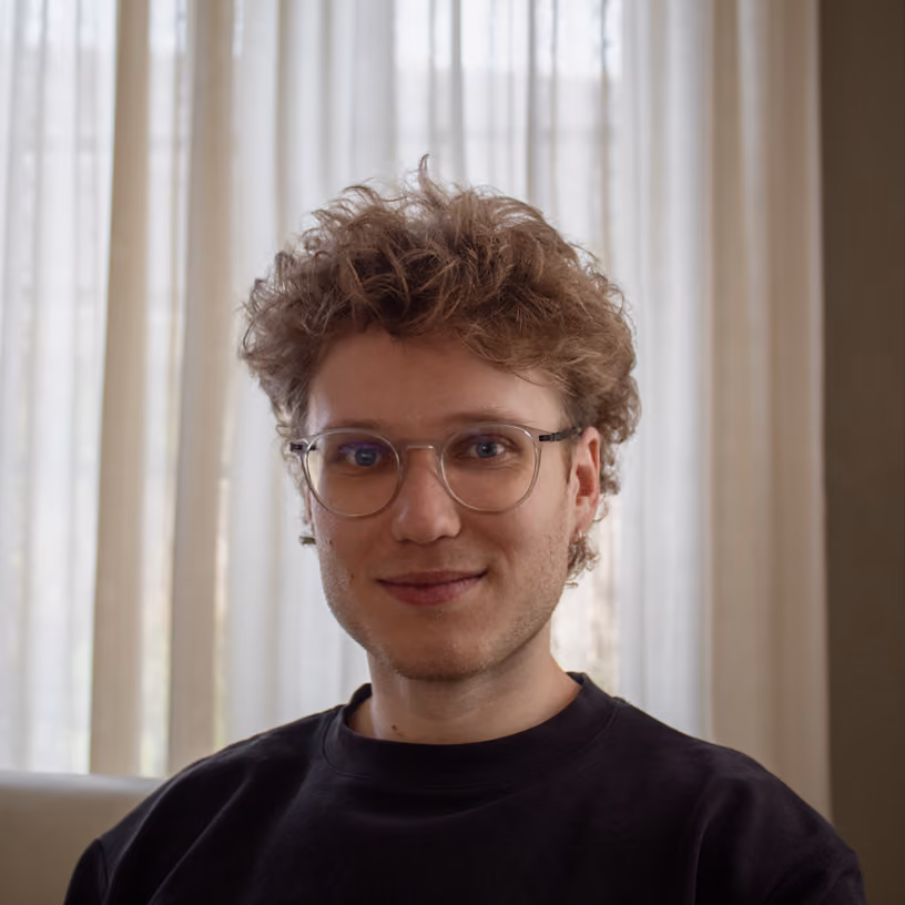 Young man with curly hair and glasses smiling while sitting indoors with sheer curtains behind him.
