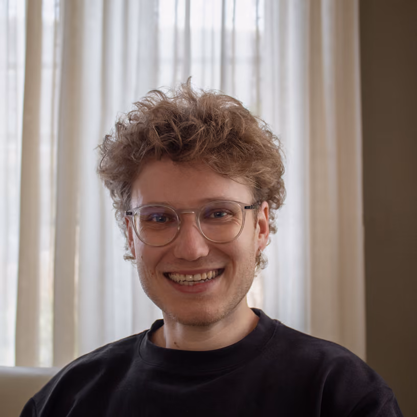 Smiling young man with curly hair and glasses wearing a black shirt in front of sheer curtains.