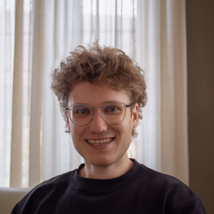 Smiling young man with curly hair and glasses wearing a black shirt, sitting indoors with curtains behind.