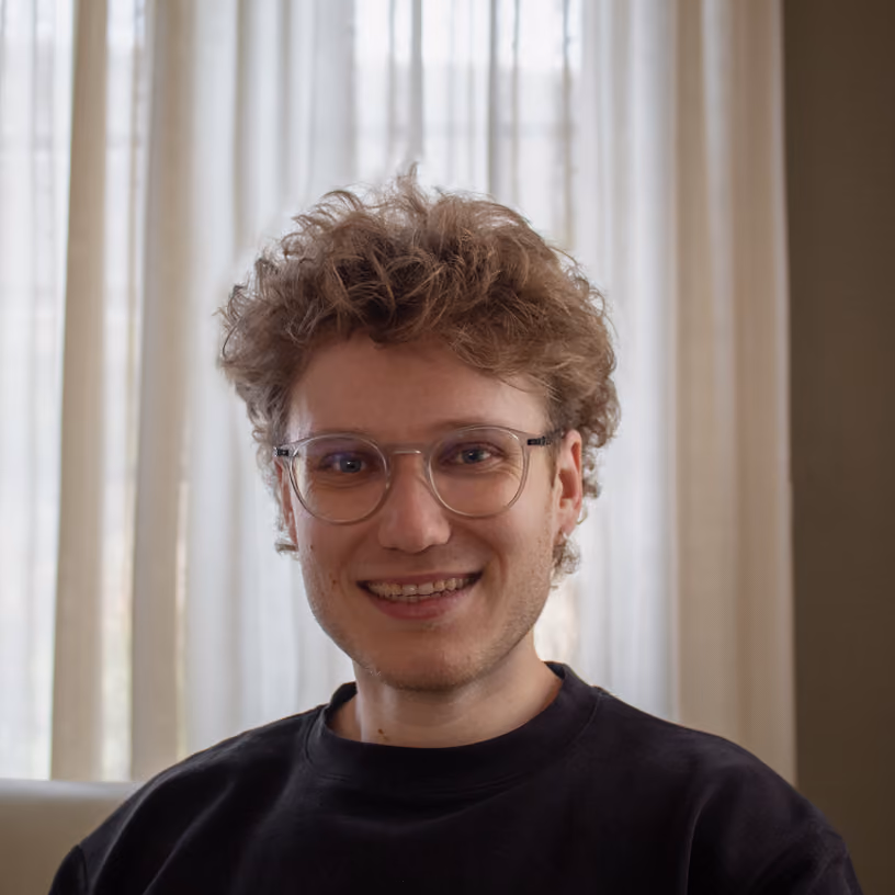 Smiling young man with curly hair and glasses wearing a black shirt sitting indoors.