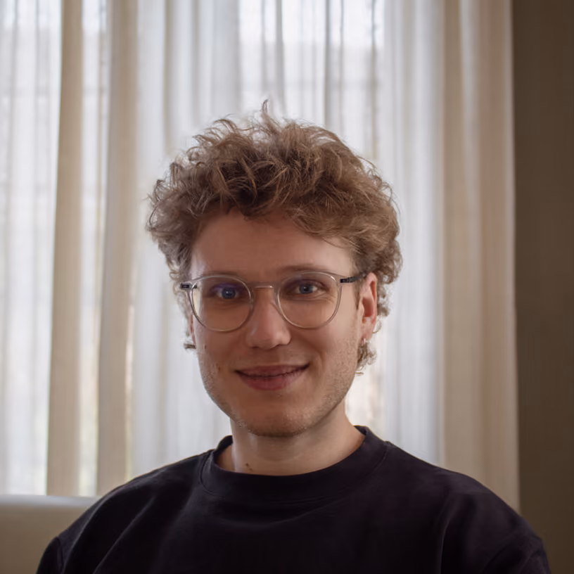 Young person with curly hair and glasses smiling in front of sheer white curtains.