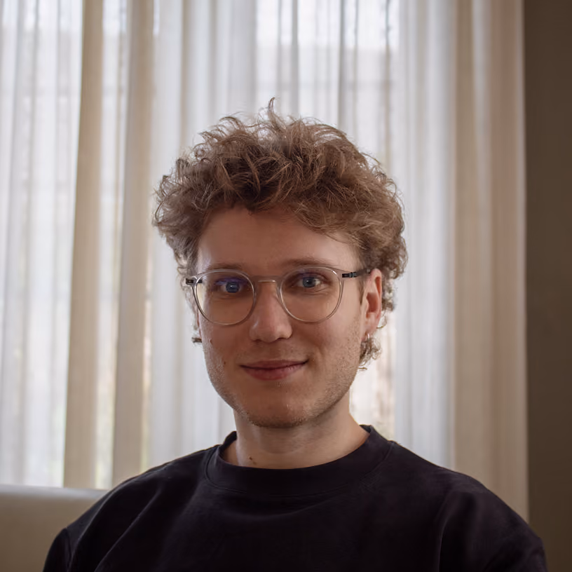 Young man with curly light brown hair and glasses wearing a dark sweater in front of sheer white curtains.
