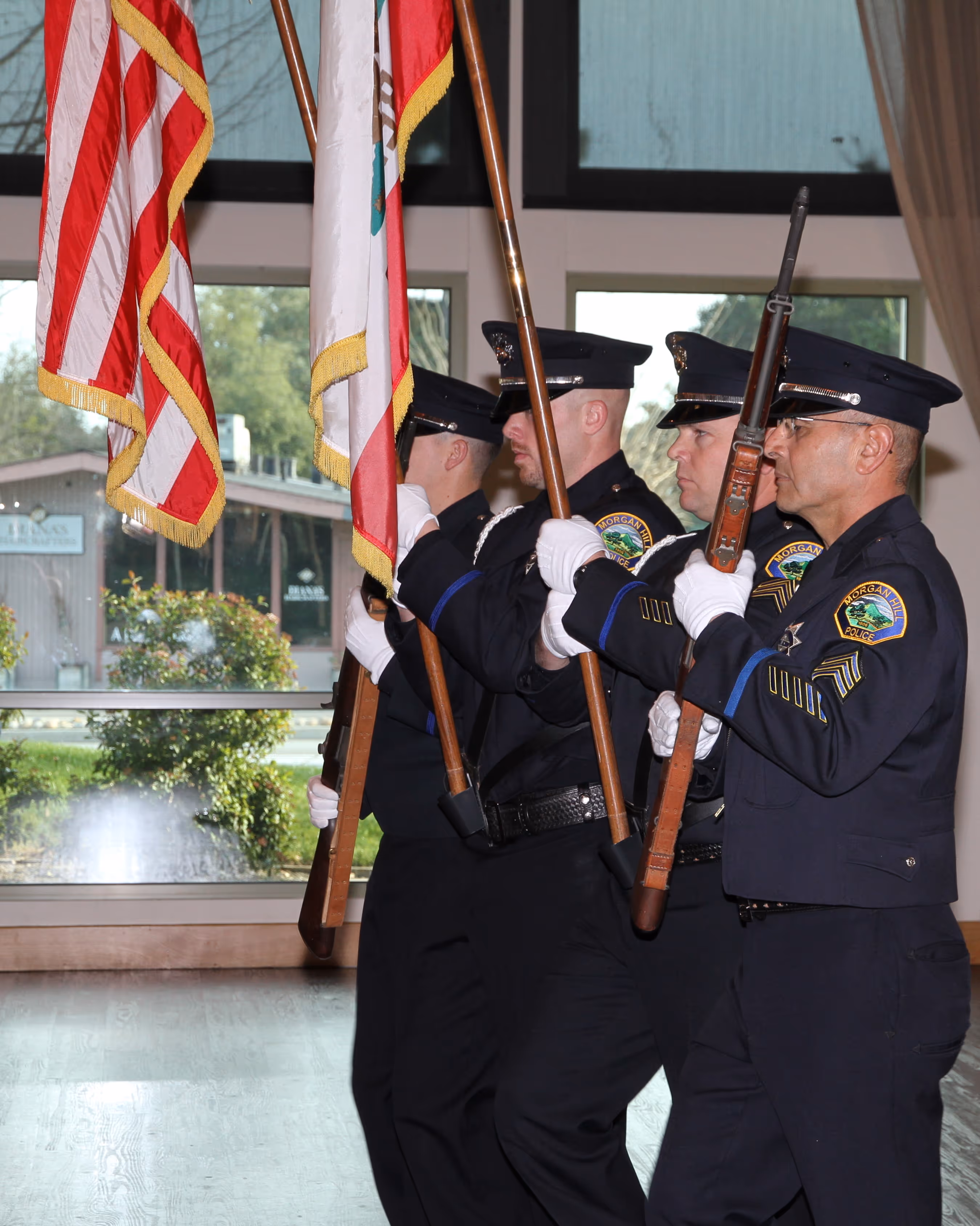Four uniformed police officers in a color guard hold flags and rifles indoors near large windows.