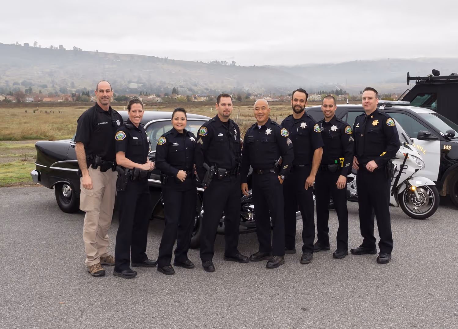 Group of eight police officers standing outdoors in front of police vehicles, posing for a photo on a cloudy day.