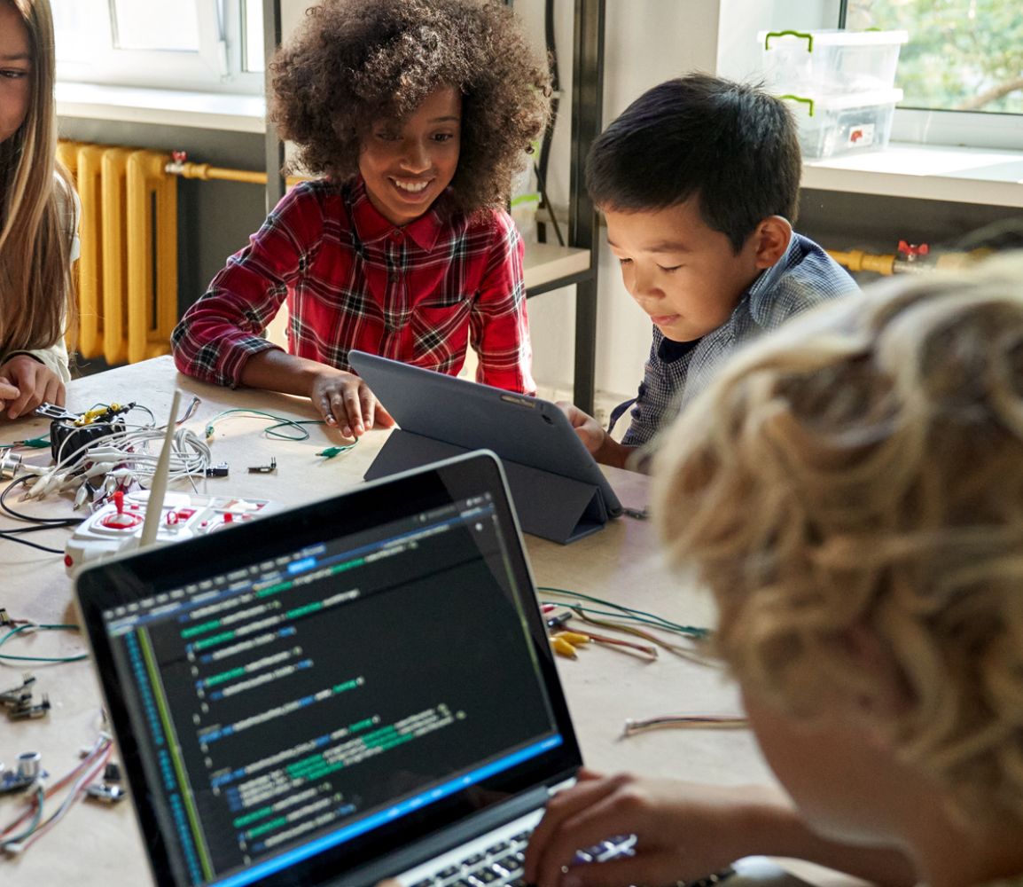 children gathered around a table with circuit parts and laptops learning to code