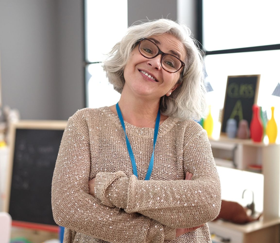 a teacher with glasses and grey hair standing with arms crossed in a classroom