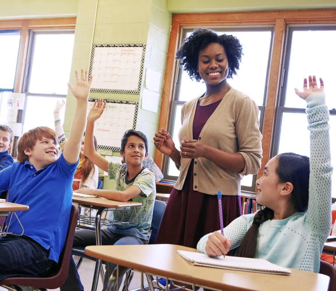 smiling teacher walking through desks of kids with hands raised