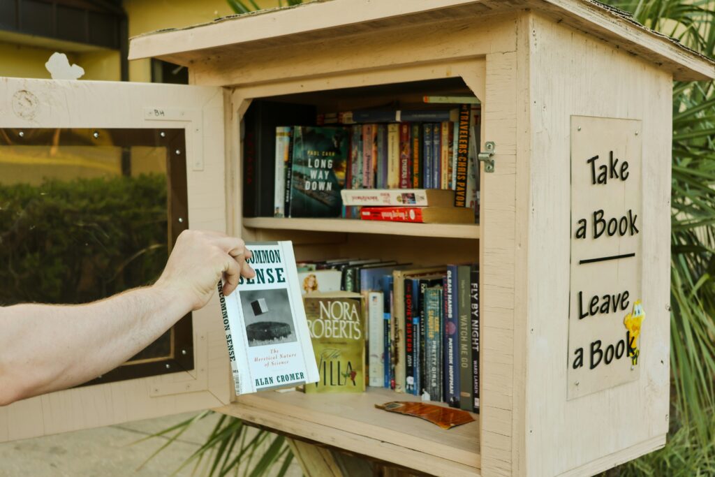 This is an Image of a free neighborhood library. The image contains a small wooden box filled with books and a person putting a new book into the library. There is writing on the side of the box that reads, "Take a Book, Leave a Book." Image by Kaylar Photo on Unsplash
