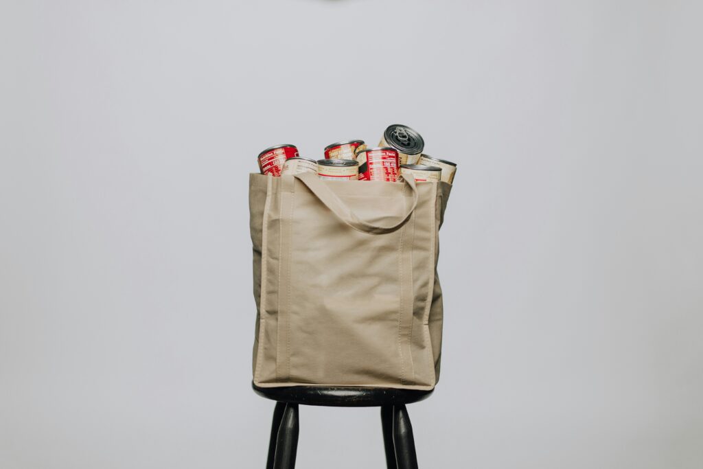 An image of a reusable bag sitting on a stool. The bag contains cans of soup with a red label. 