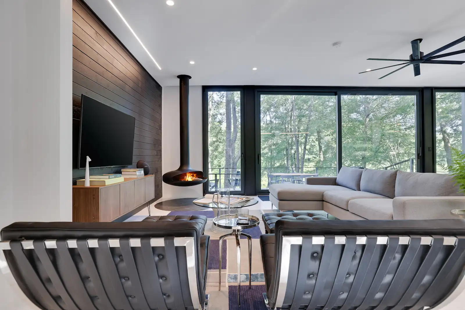 Modern living room with black chairs, beige sofa, hanging fireplace, and large glass windows showing trees.