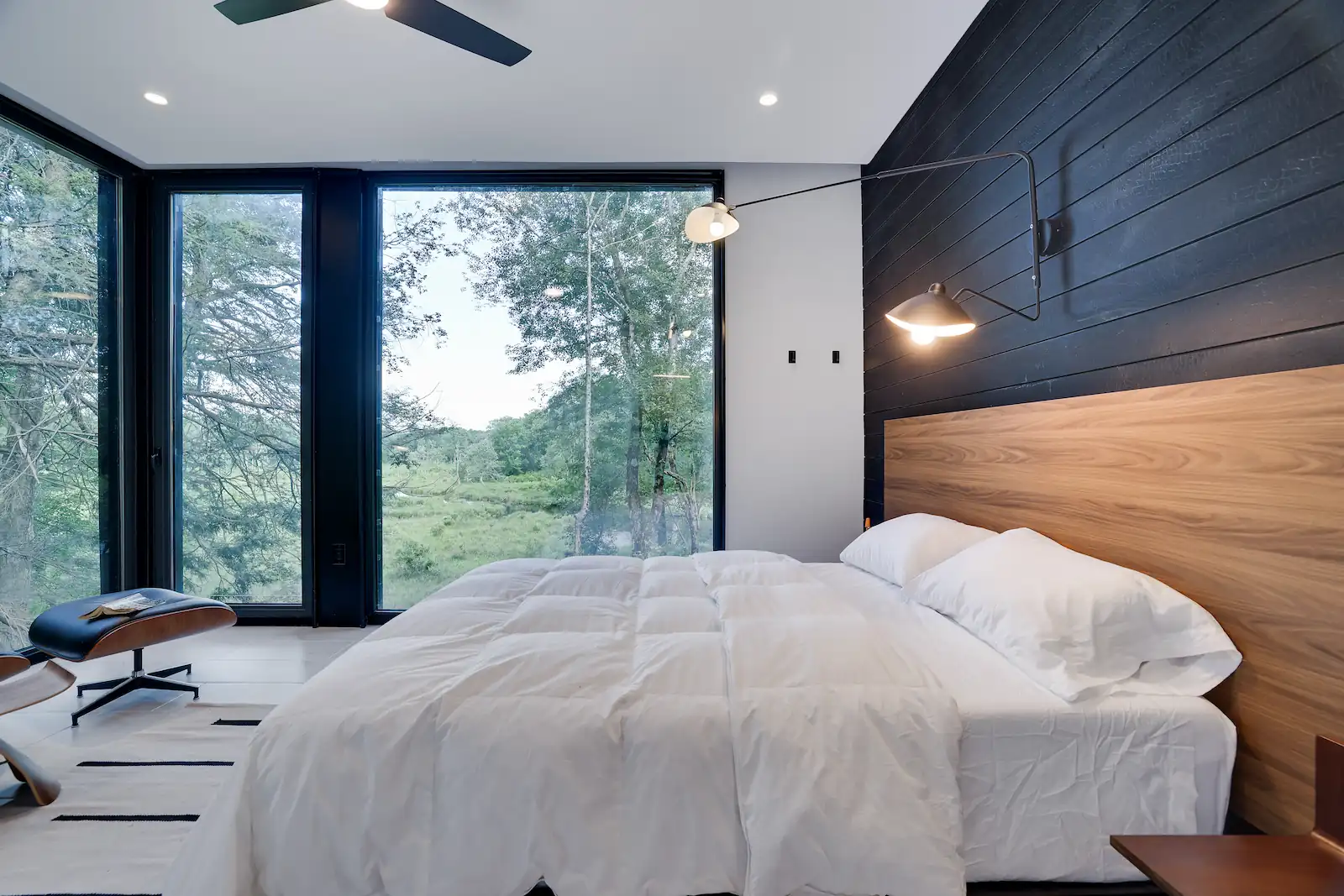 Modern bedroom with a wood-paneled headboard wall, wall-mounted reading lights, and floor-to-ceiling windows facing the forest
