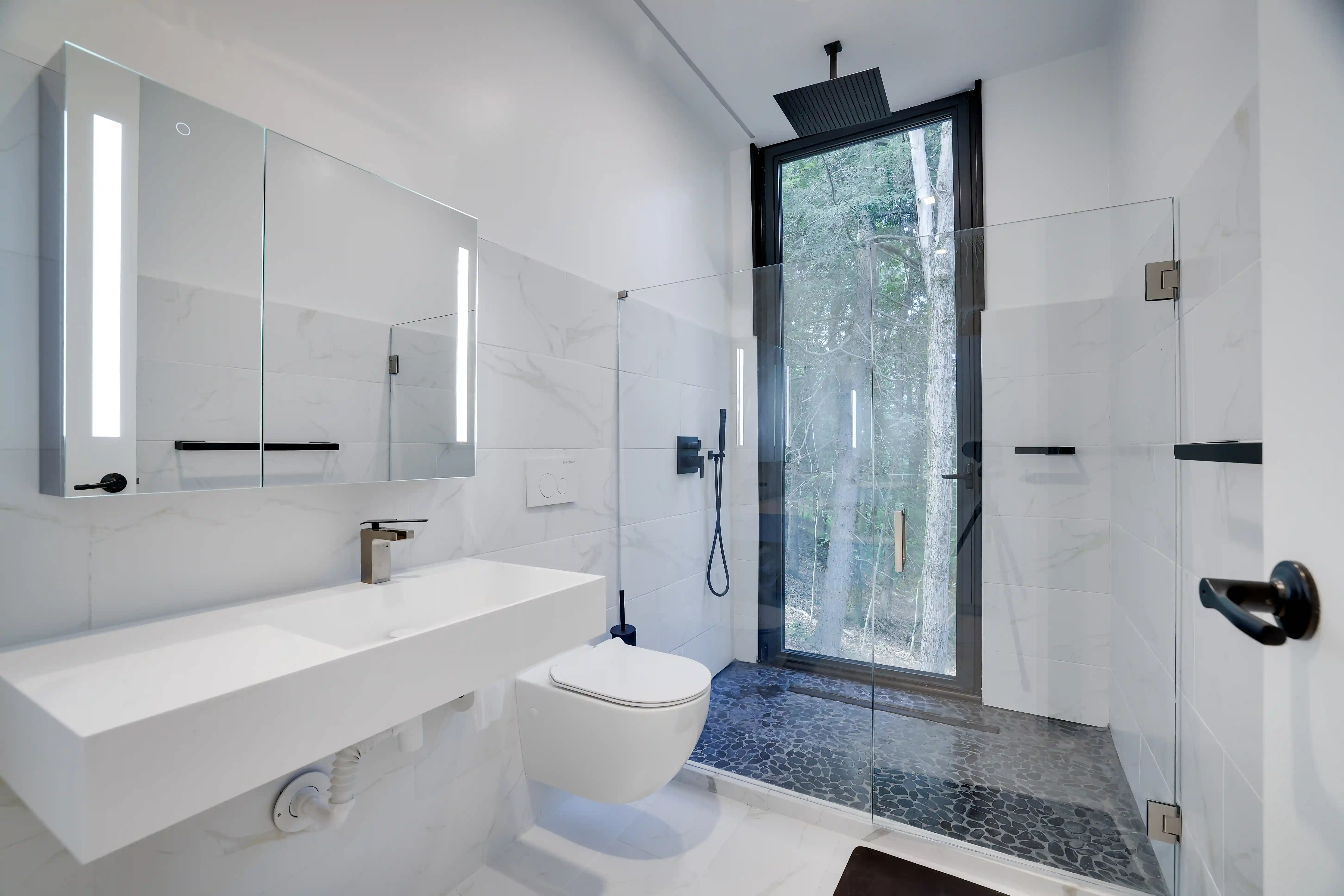 Modern white bathroom with a floating sink, mirrored medicine cabinet, and a glass-walled shower with a forest view