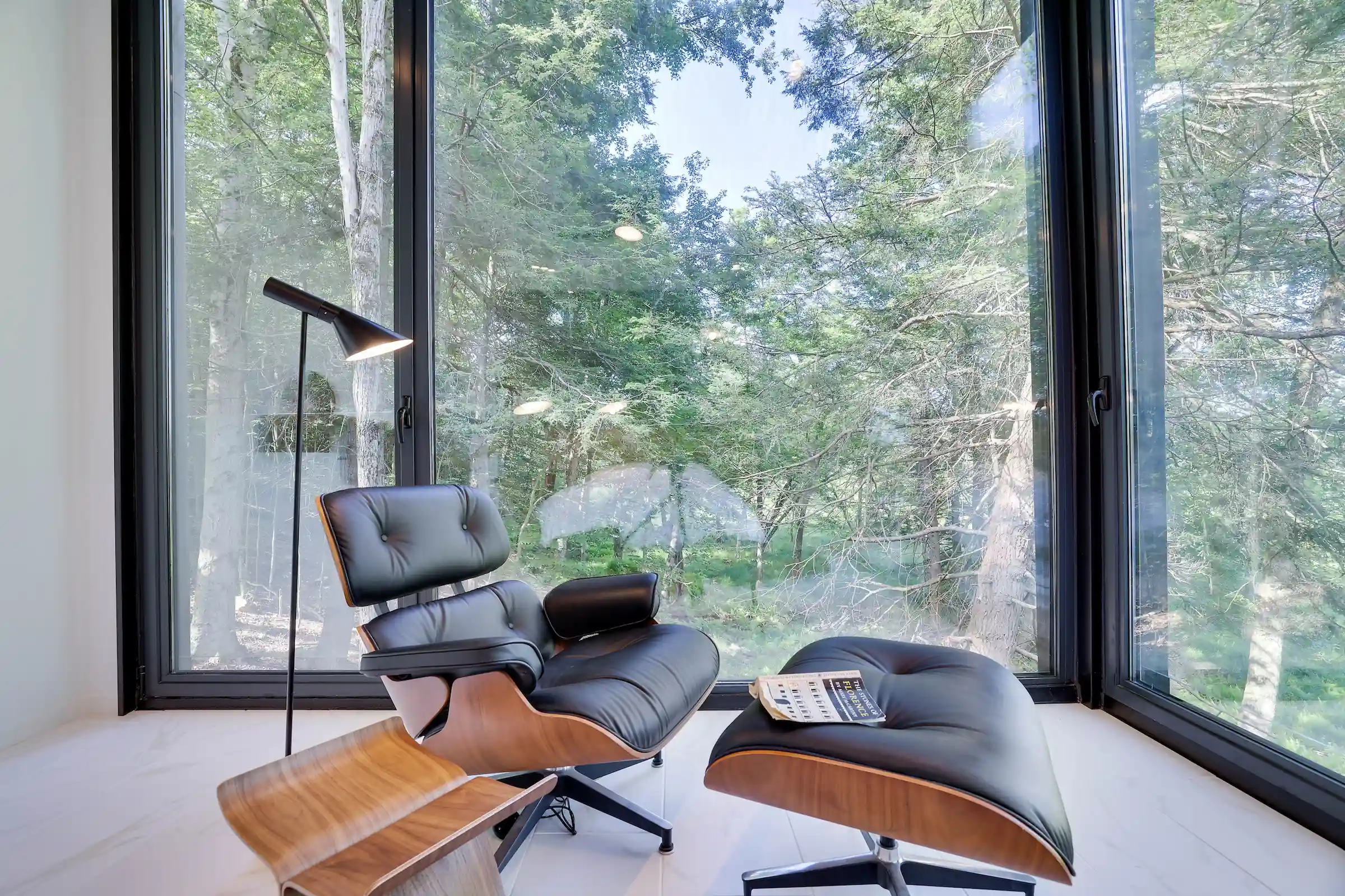 Reading nook with a black leather Eames lounge chair and ottoman beside floor-to-ceiling windows overlooking the forest