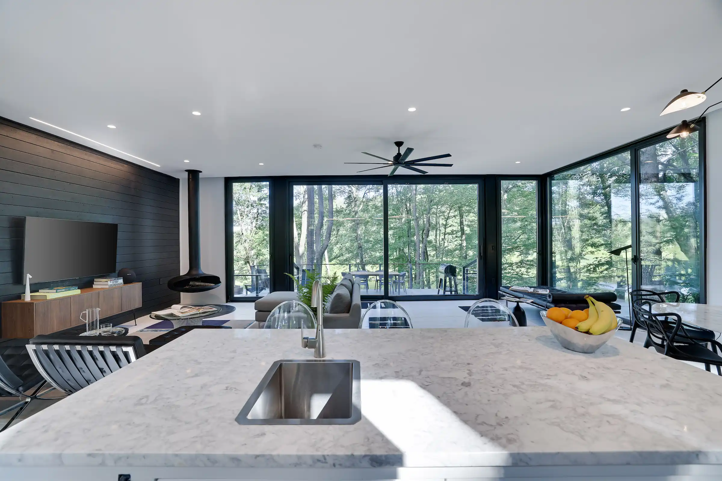 Open-plan living area with a black ceiling fan, floor-to-ceiling windows overlooking the forest, marble kitchen island, and dining table