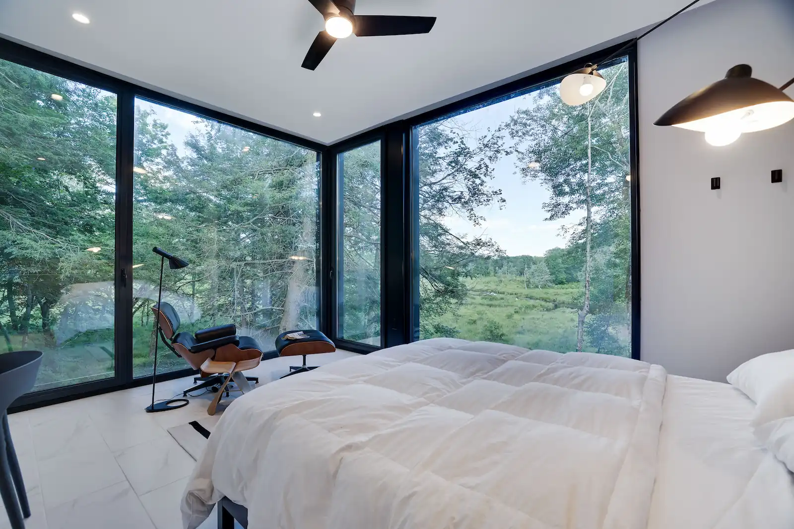 Bedroom corner with an Eames lounge chair facing floor-to-ceiling glass overlooking the meadow and forest.