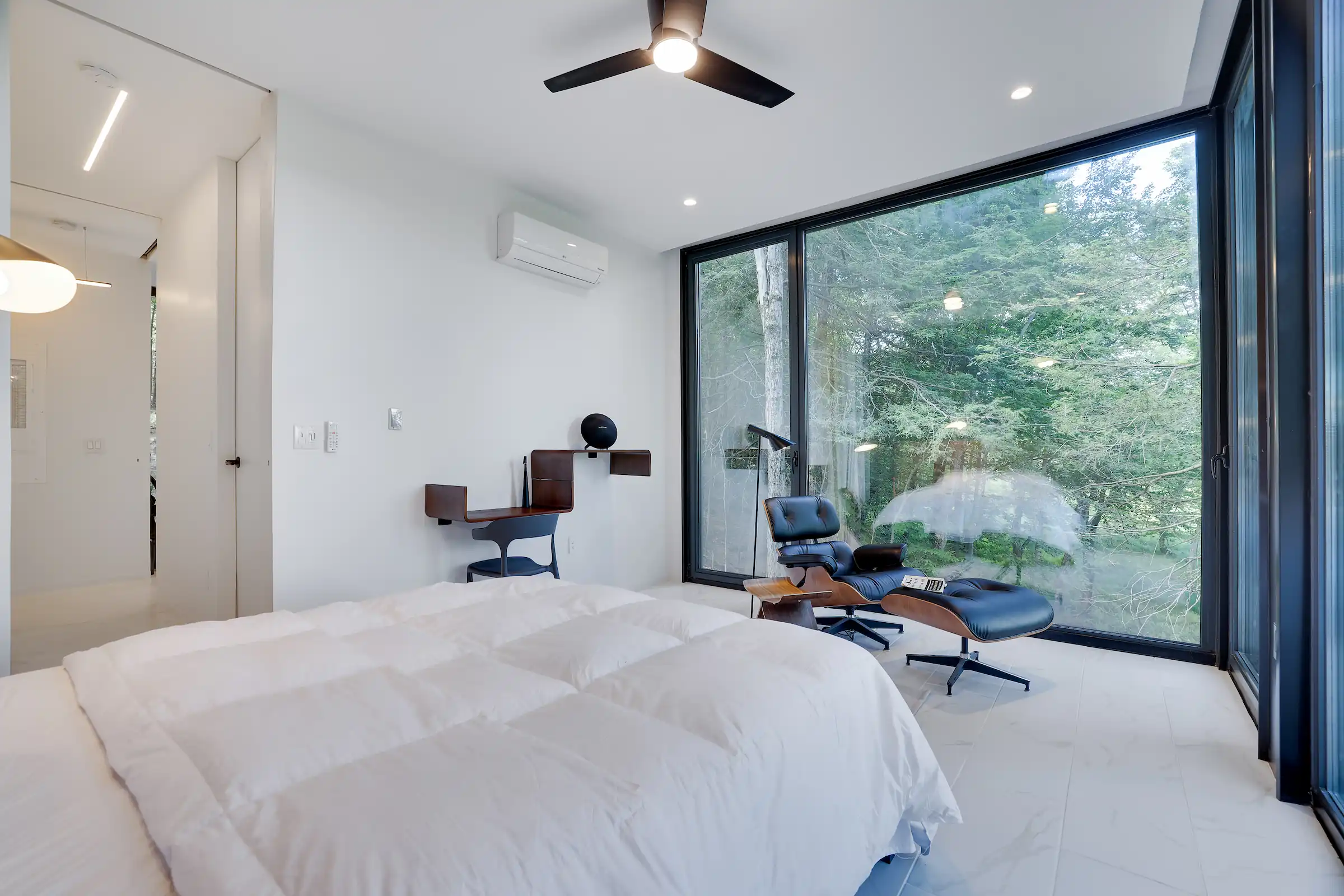 Bedroom with a corner desk nook and Eames lounge chair beside glass walls framing the trees.