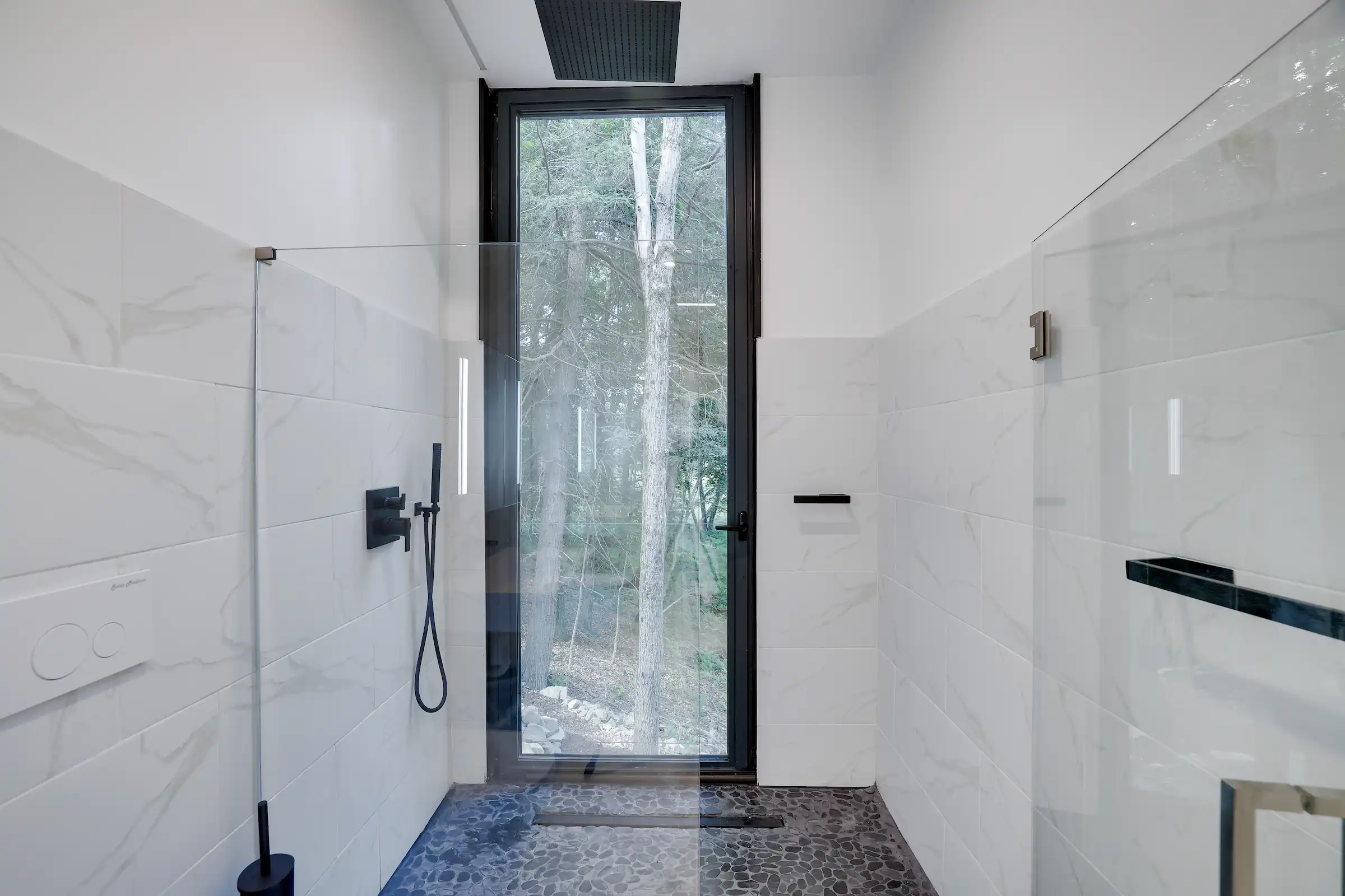 Marble-tiled walk-in shower with black fixtures and a tall window overlooking the forest.