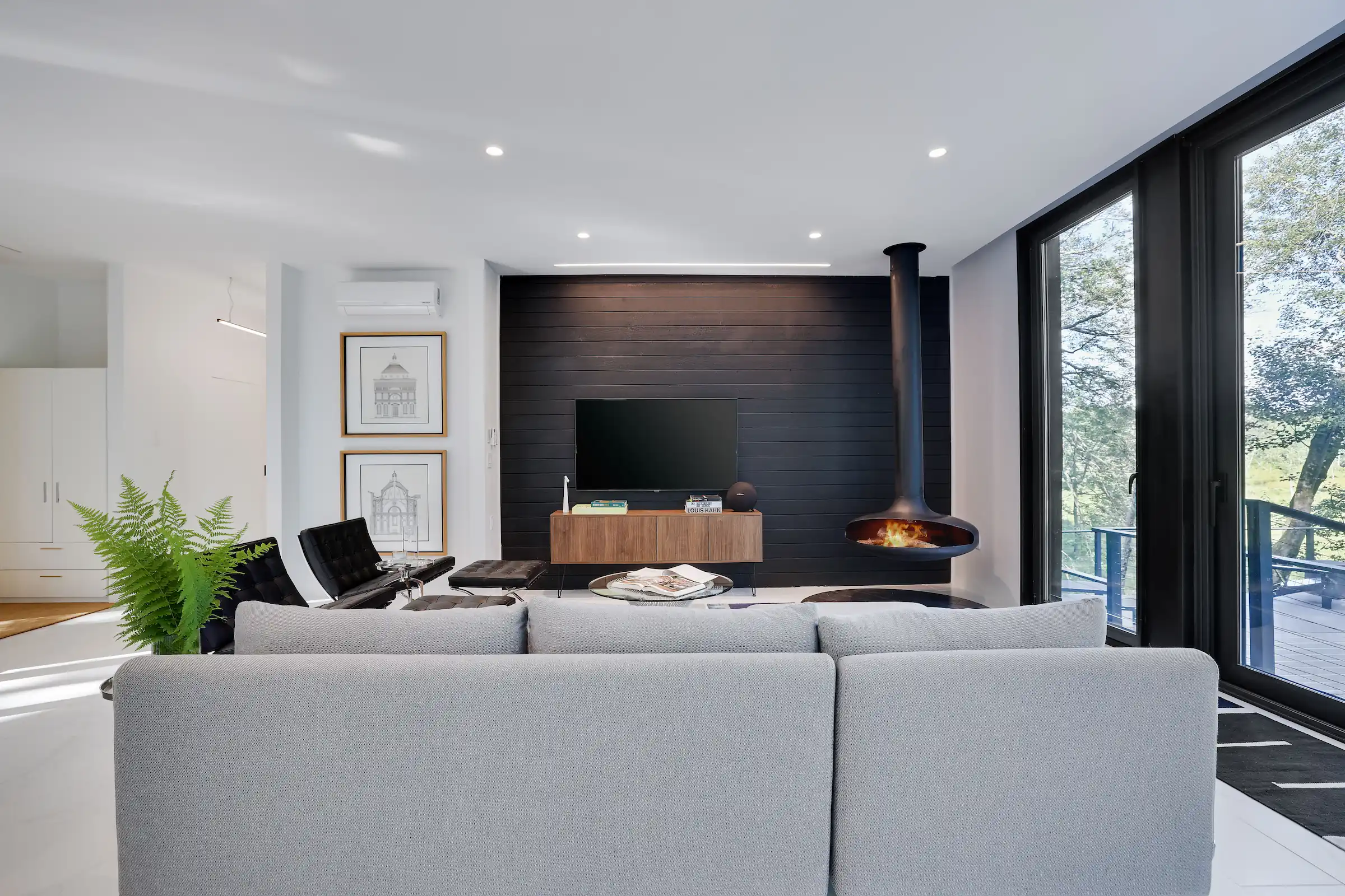 Gray sectional sofa facing a charred wood accent wall with a suspended fireplace and floor-to-ceiling glass doors opening to the forest.