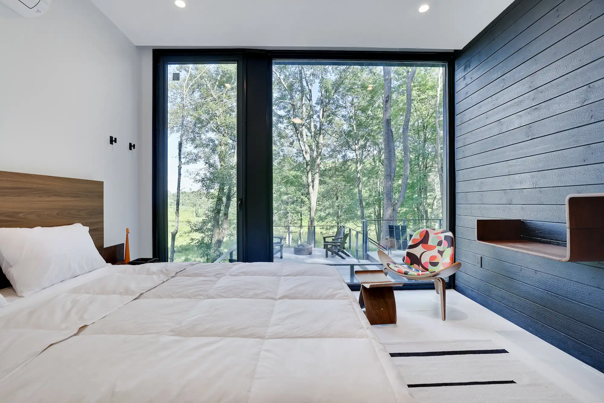 Bedroom with charred wood accent wall and a corner glass window overlooking the trees.