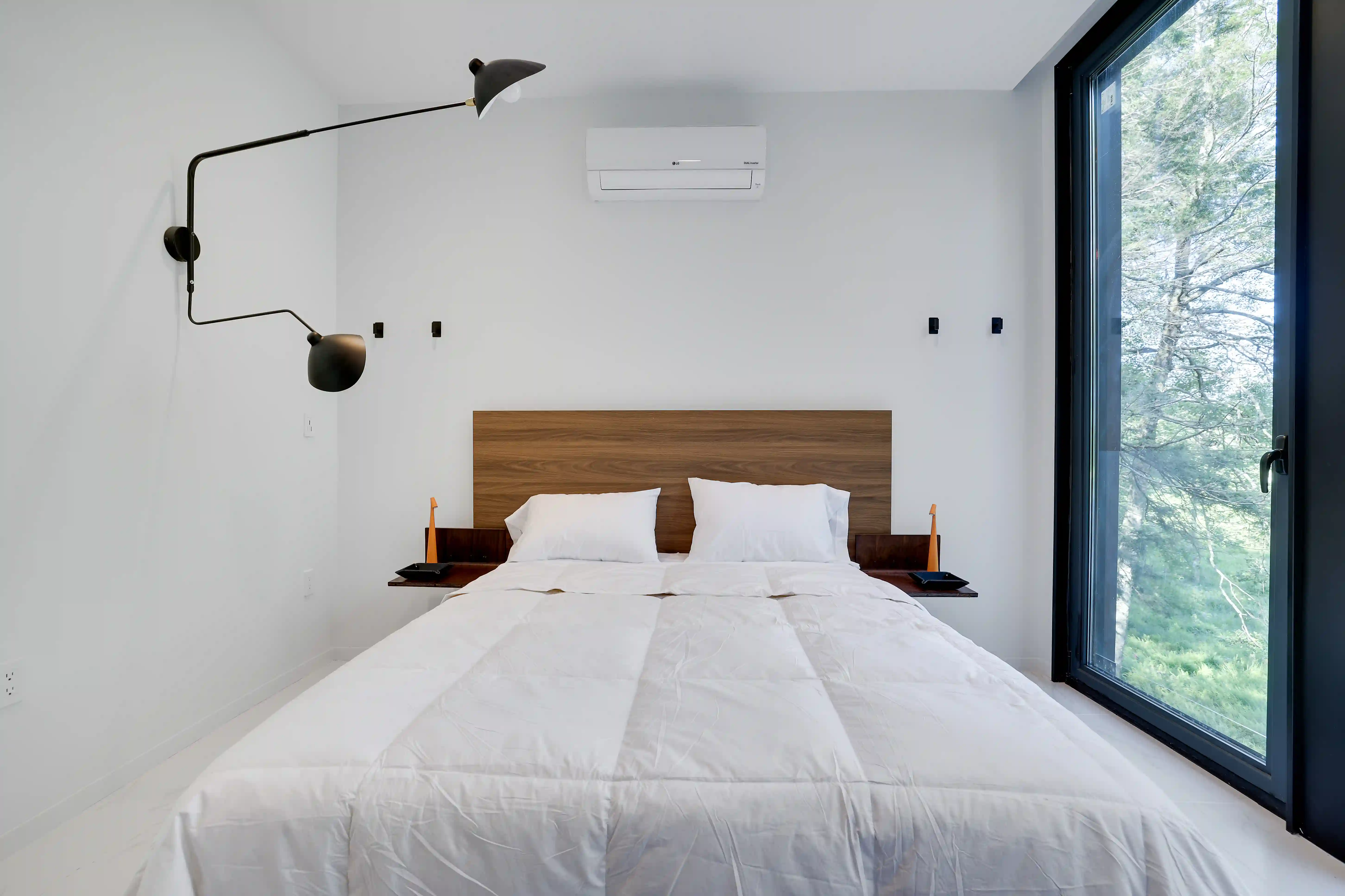 Walnut headboard bed with Egyptian cotton linens beneath a sconce, lit by a window view of the forest.