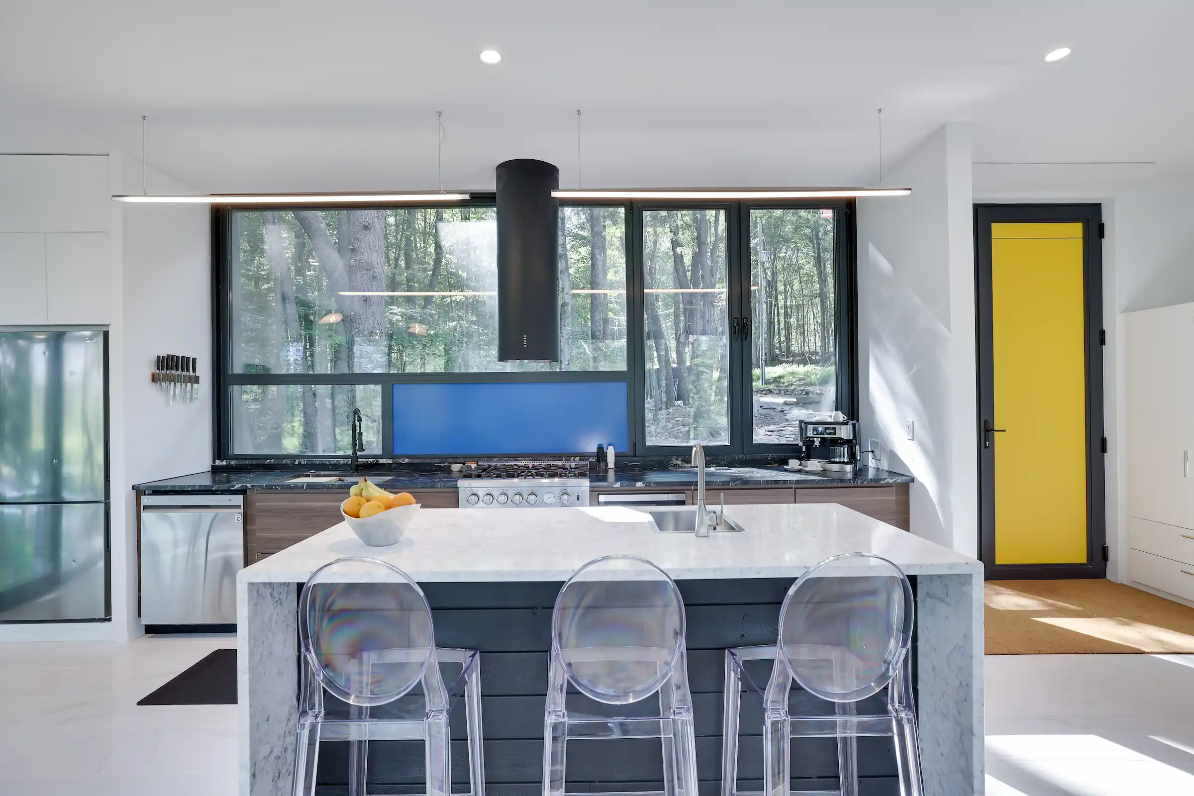 Waterfall marble kitchen island with ghost stools, gas range, and a forest-view window backsplash.