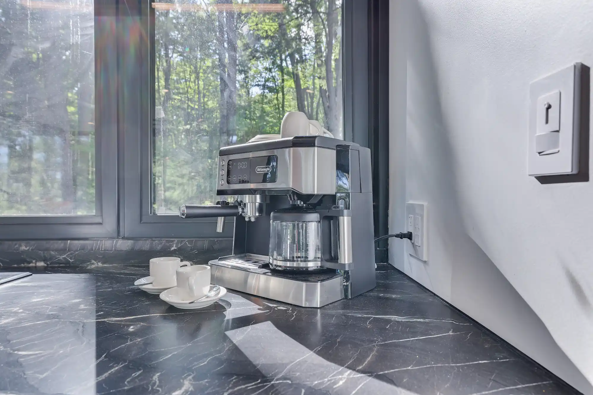Espresso machine and cups set on a dark marble counter beside a forest-view window.